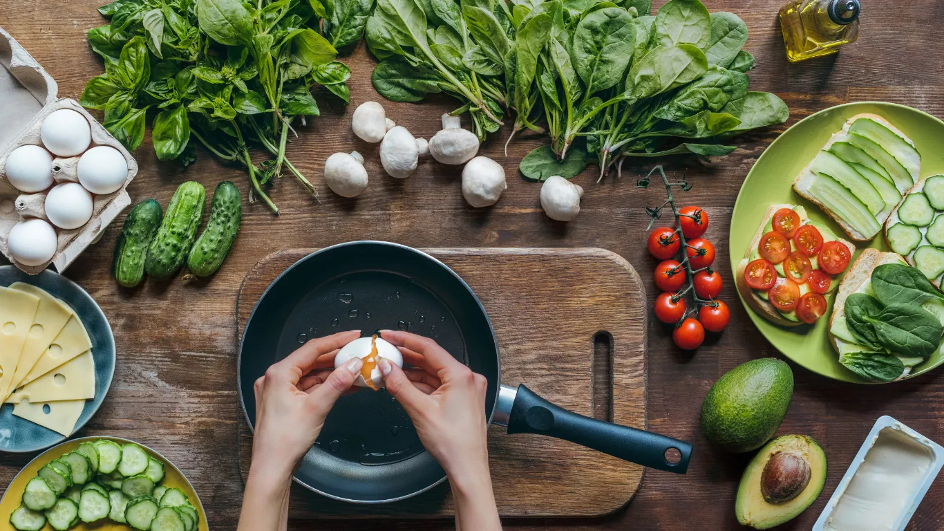 Hands cracking an egg into a pan, with fresh ingredients and a phone showing the Pantry Pilot app nearby.